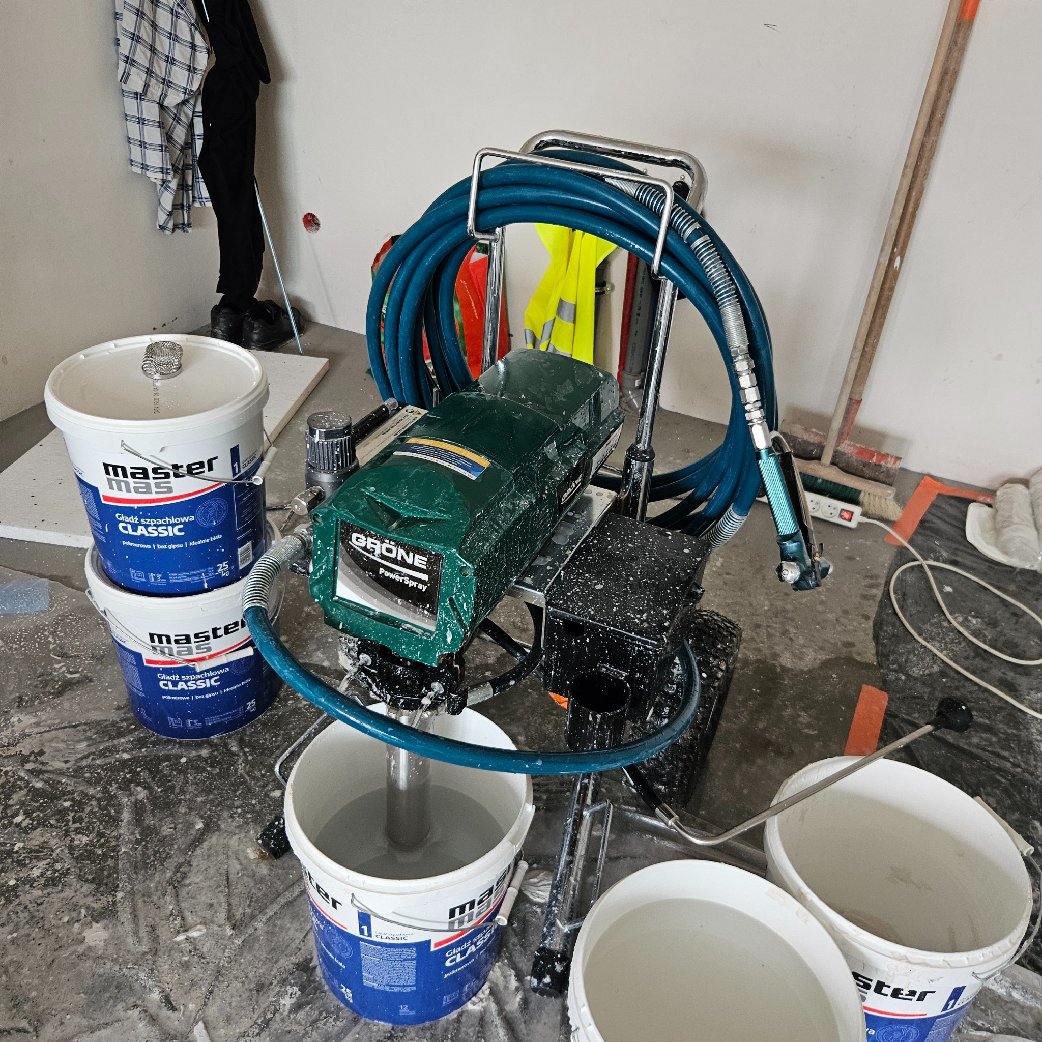Agregat malarski Grone PowerSpray z niebieskim wężem i kilkoma wiadrami z gładzią szpachlową Master Mas Classic na folii ochronnej w trakcie prac wykończeniowych.