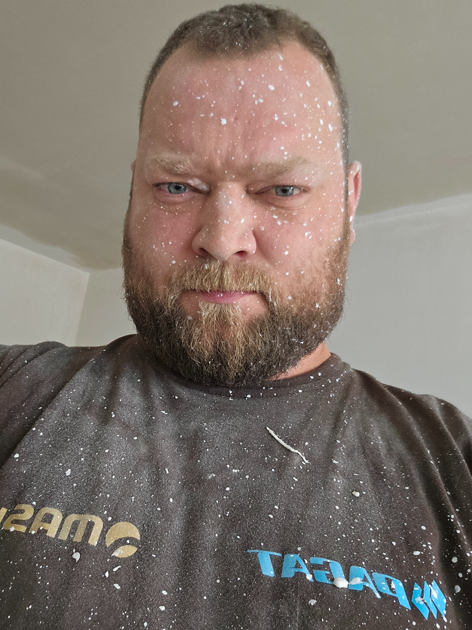 Mężczyzna z brodą, pokryty białymi plamami farby lub gładzi, spogląda z góry, ubrany w brudną koszulkę z logo firmy budowlanej.