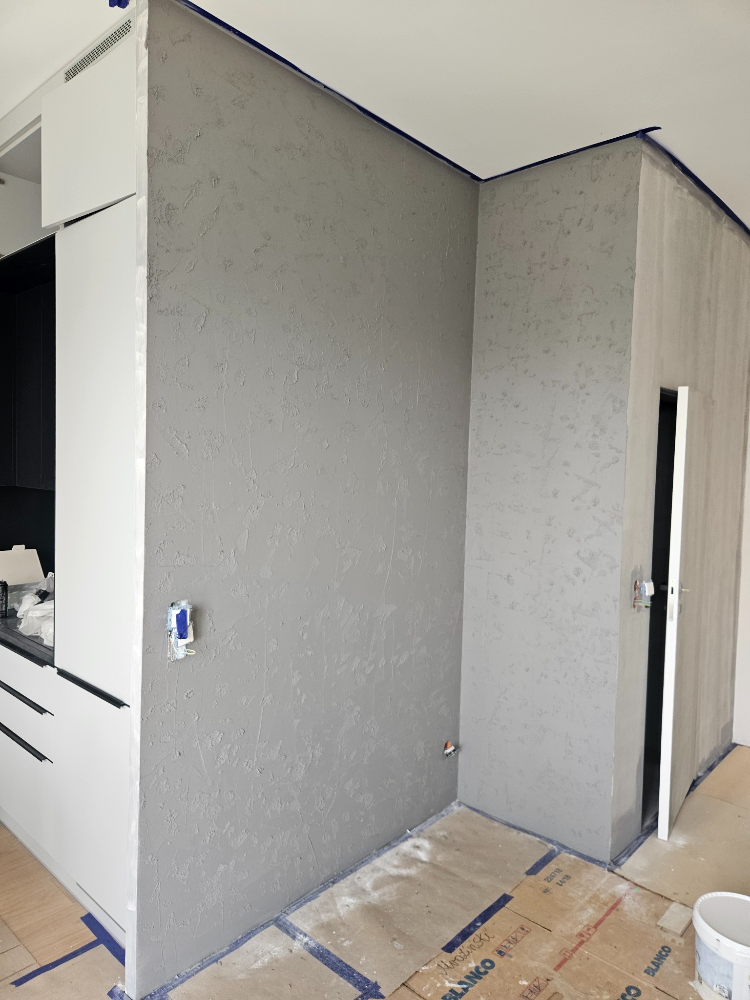 Świeżo położona, szara struktura na ścianie w rogu pomieszczenia, zabezpieczona niebieską taśmą malarską przy suficie i podłodze, z widoczną białą zabudową kuchenną po lewej stronie i otwartymi...