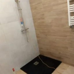 Niedokończona łazienka z czarnym brodzikiem, jasnymi płytkami na ścianach i grzejnikiem drabinkowym. Widoczny przewód i butelka z płynem do czyszczenia.