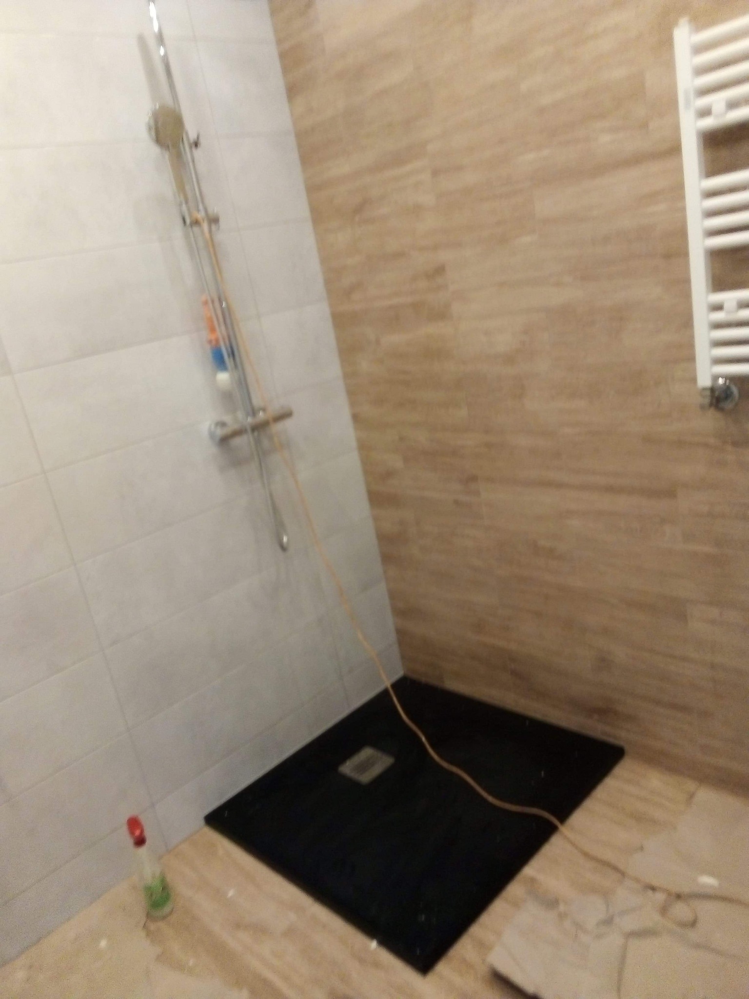 Niedokończona łazienka z czarnym brodzikiem, jasnymi płytkami na ścianach i grzejnikiem drabinkowym. Widoczny przewód i butelka z płynem do czyszczenia.