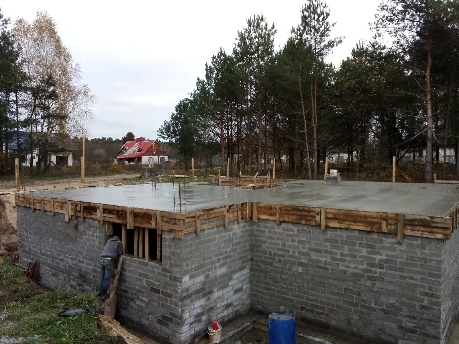 Wylany fundament budynku z szalunkiem i wystającymi prętami zbrojeniowymi, pracownik wchodzi po drabinie do wnętrza fundamentu, w tle las i dom w trakcie remontu.