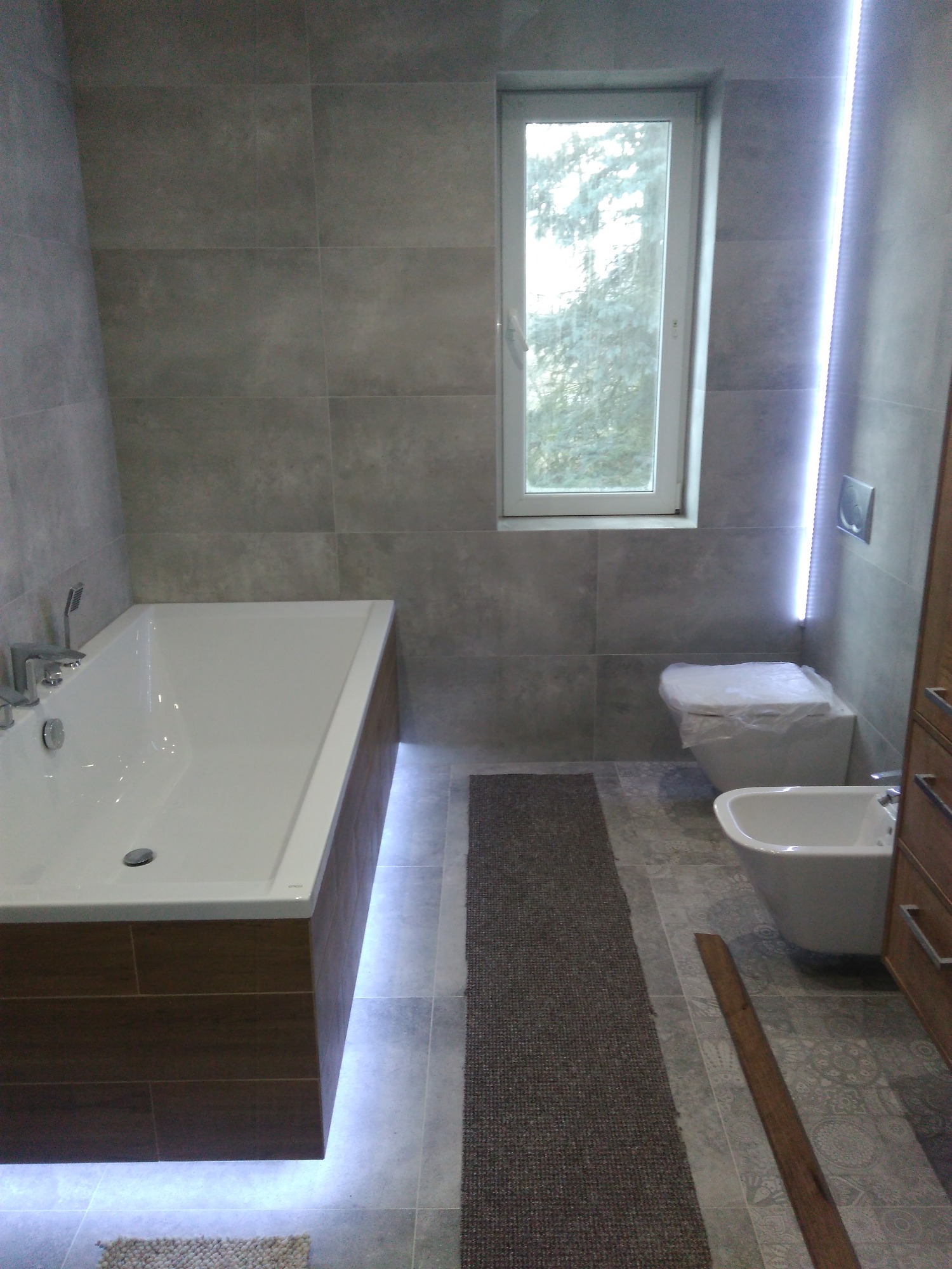 Nowoczesna łazienka z szarymi płytkami, białą wanną z podświetleniem LED, wiszącą toaletą w folii ochronnej, bidetem i drewnianą szafką, uzupełniona o dywanik i oświetlenie LED wzdłuż ściany.