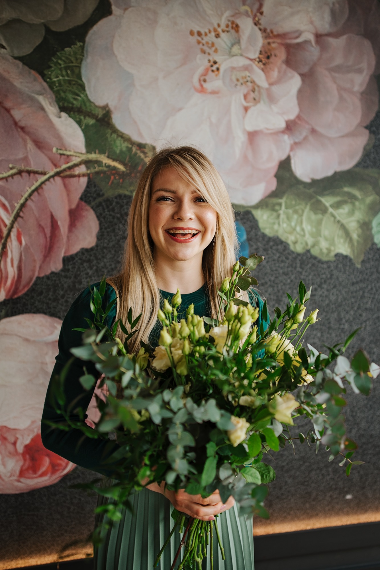 Uśmiechnięta blondynka w zielonej sukience trzyma bukiet kremowych róż i eukaliptusa na tle ściany z motywem kwiatowym.