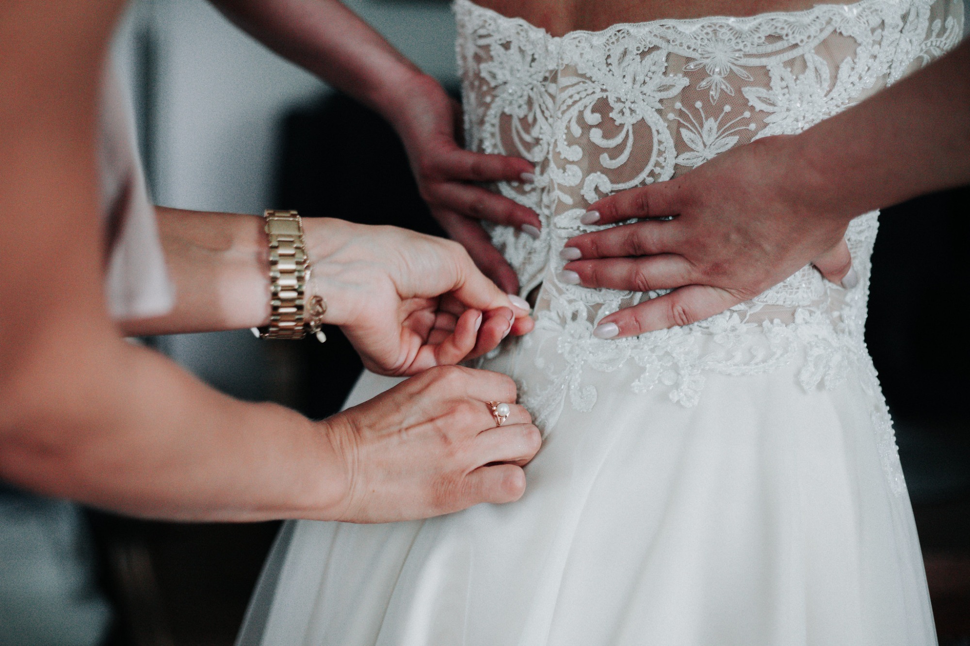 Zapinanie sukni ślubnej z koronkowym gorsetem, widoczny złoty zegarek i pierścionek z perłą na dłoniach.