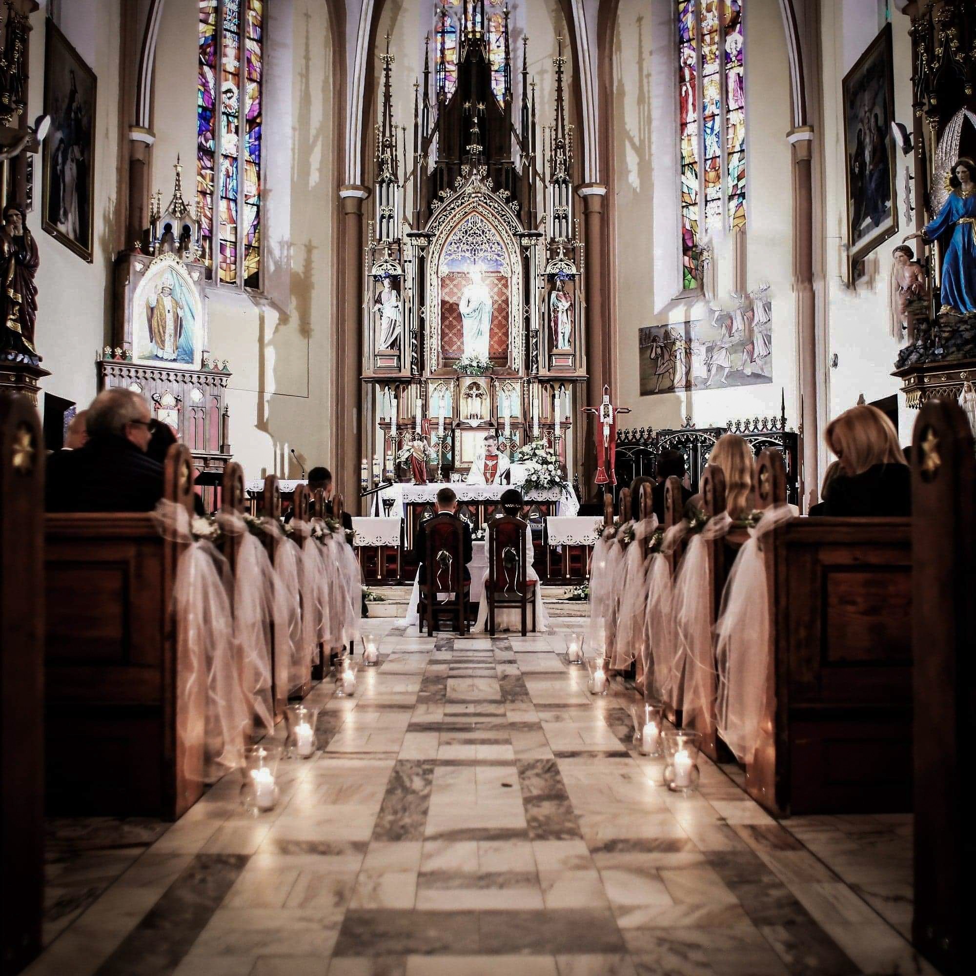 Wnętrze kościoła podczas ceremonii ślubnej, widok na parę młodą siedzącą przed ołtarzem, ławki udekorowane białymi tiulami i świecami.