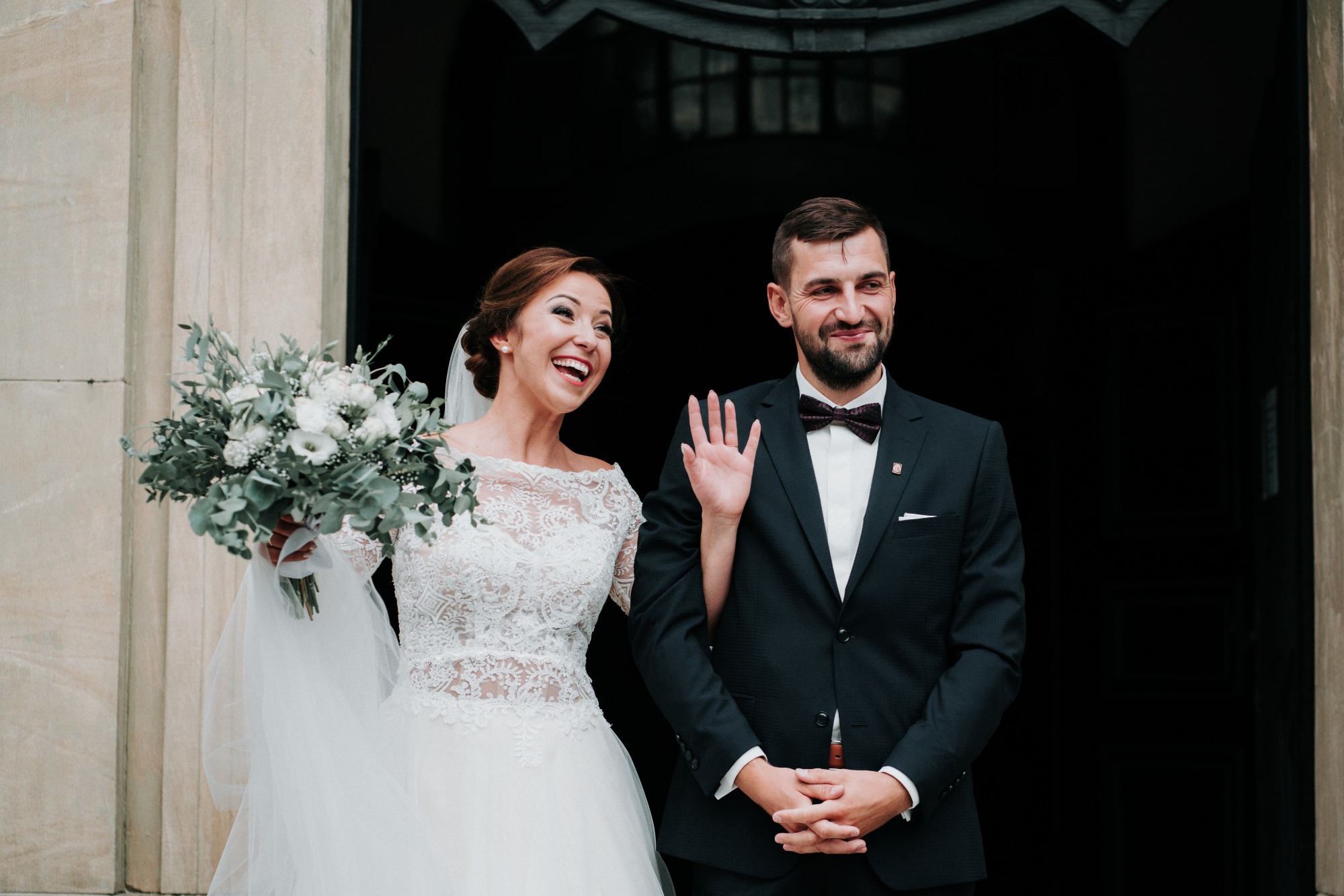 Uśmiechnięta panna młoda w koronkowej sukni z bukietem i pan młody w ciemnym garniturze z bordową muchą, stojący przed ciemnym wejściem.