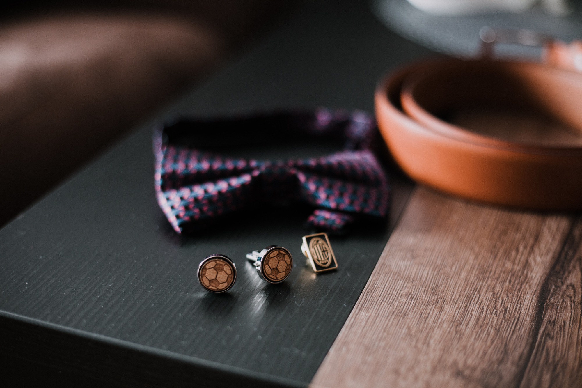 Męskie akcesoria we Wrocławiu: muszka w bordowo-granatową kratę, spinki do mankietów z motywem piłki nożnej i przypinka na ciemnym blacie z elementami drewna, skórzany pasek w tle.