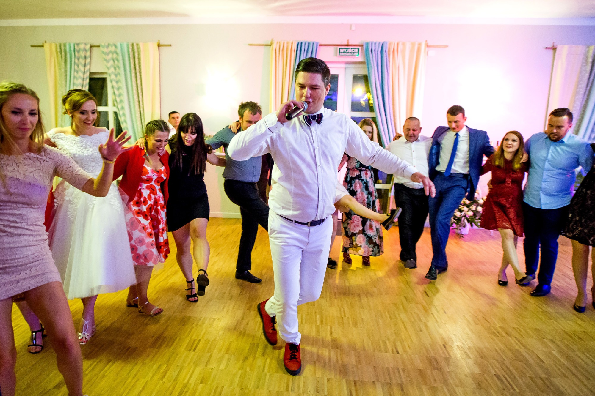 Rozbawiony wodzirej w białym garniturze i czerwonych butach prowadzi taniec weselny z mikrofonem w dłoni, otoczony przez gości w różnorodnych strojach na parkiecie.