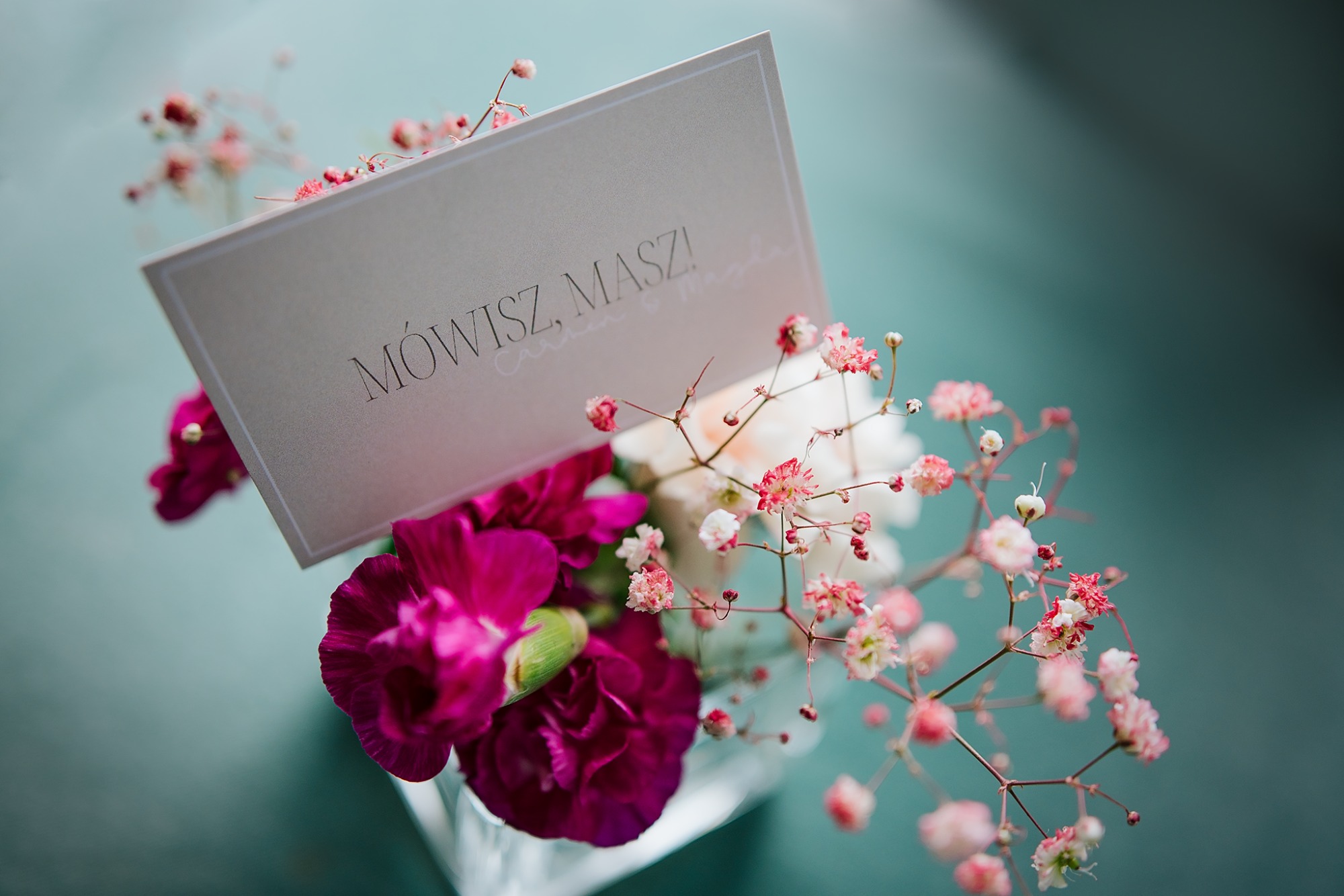 Kompozycja kwiatowa z ciemnoróżowych goździków i gipsówki, z kartką z napisem 'Mówisz, Masz!'