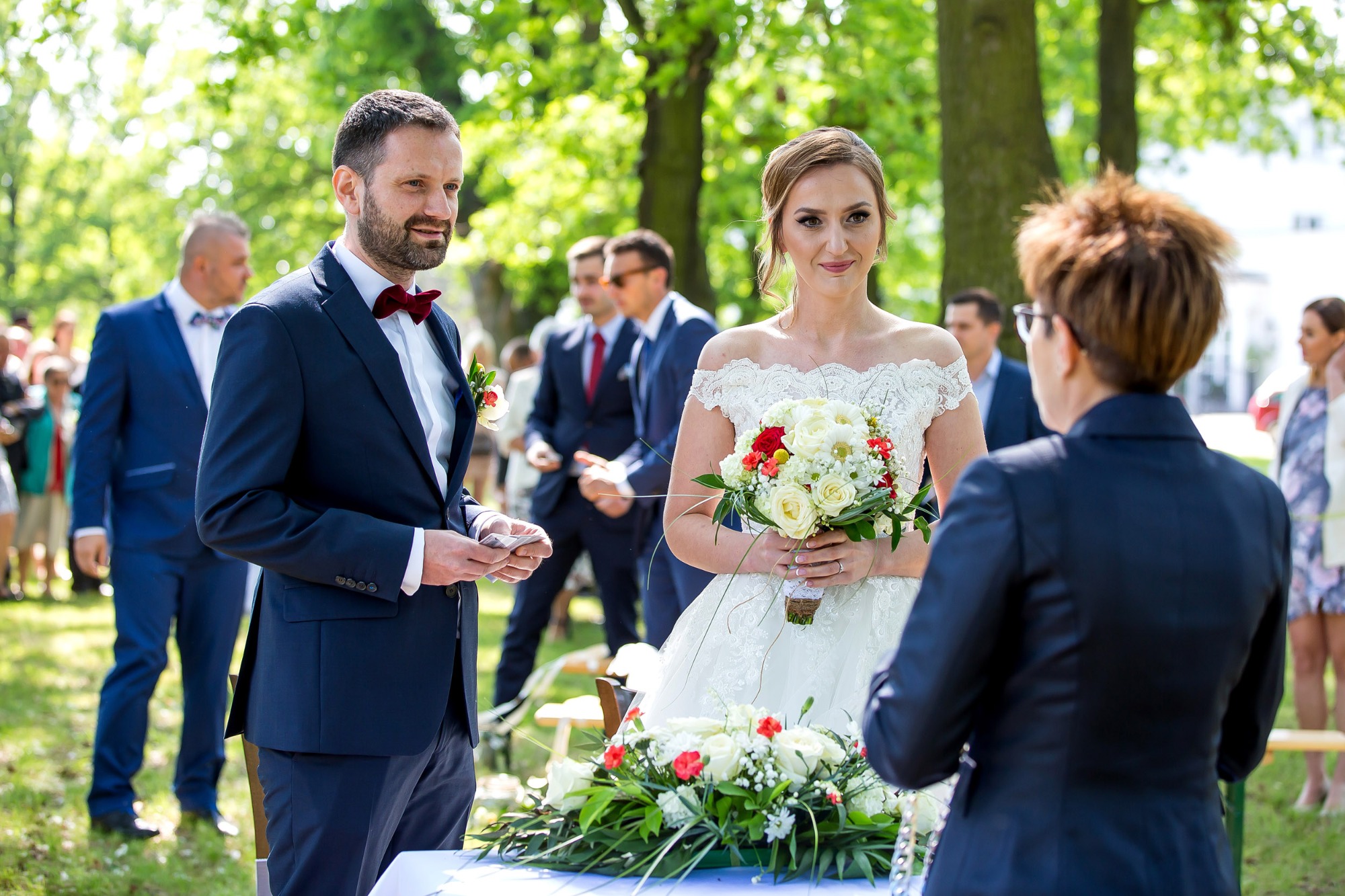 Para młoda podczas ceremonii ślubnej w plenerze, panna młoda trzyma bukiet białych i czerwonych róż, pan młody w granatowym garniturze z bordową muchą.