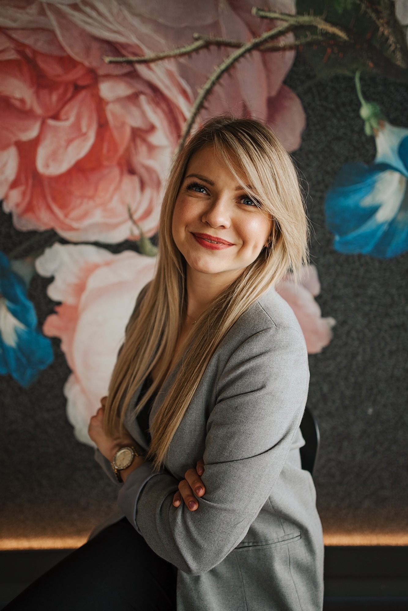 Portret uśmiechniętej blondynki w szarym żakiecie, z założonymi rękami na tle ściany z motywem kwiatowym w odcieniach różu i błękitu.