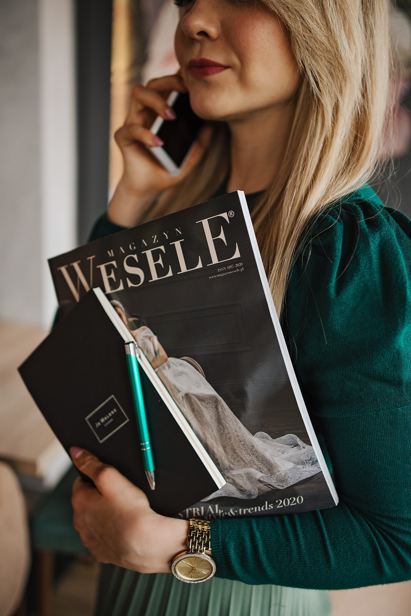 Kobieta z blond włosami rozmawia przez telefon, trzymając magazyn 'Wesele' i notatnik Jo Malone z zielonym długopisem, w tle zielona sukienka i złoty zegarek.