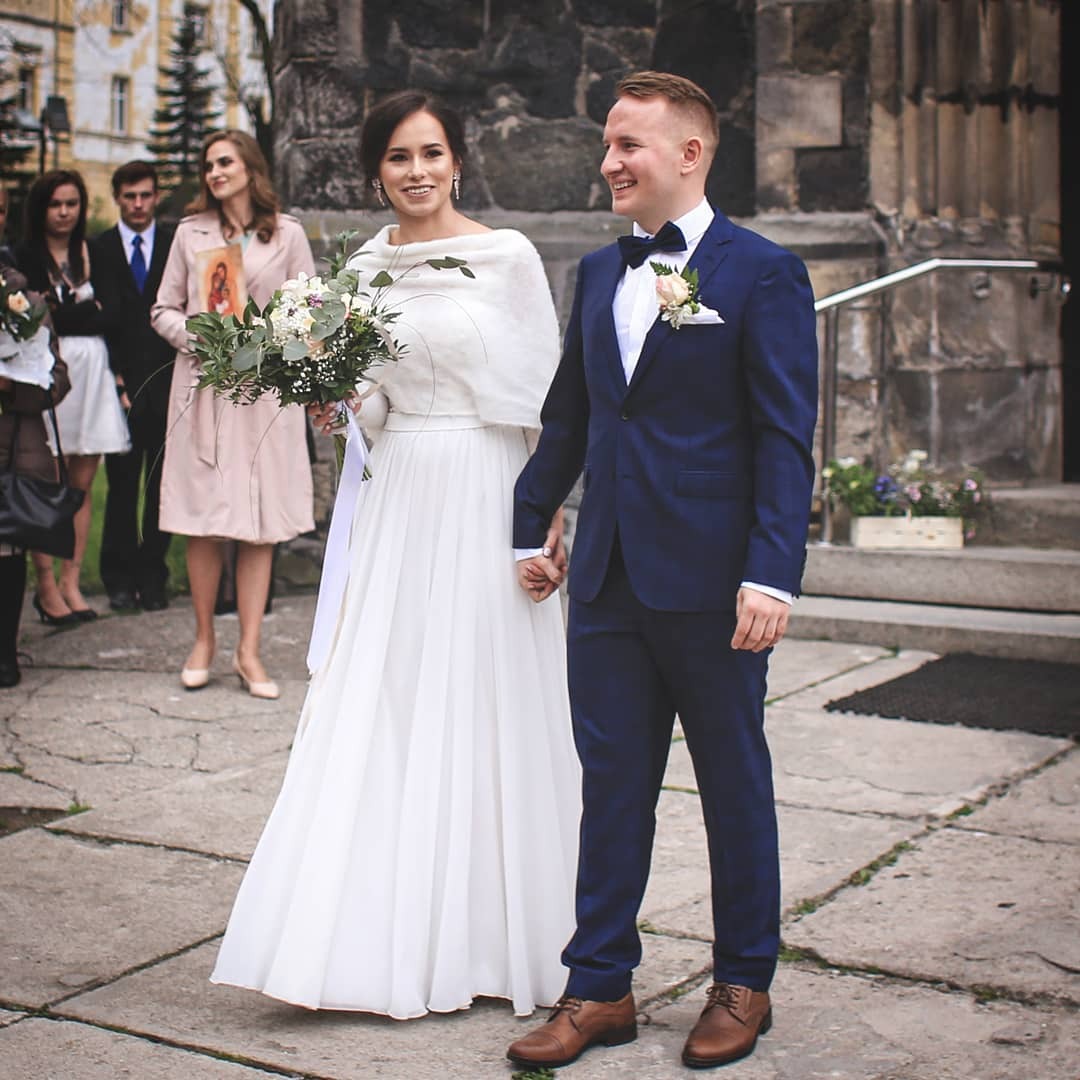 Para młoda, ona w białej sukni i z bukietem, on w granatowym garniturze, trzymają się za ręce na tle kamiennego budynku, goście weselni w tle.