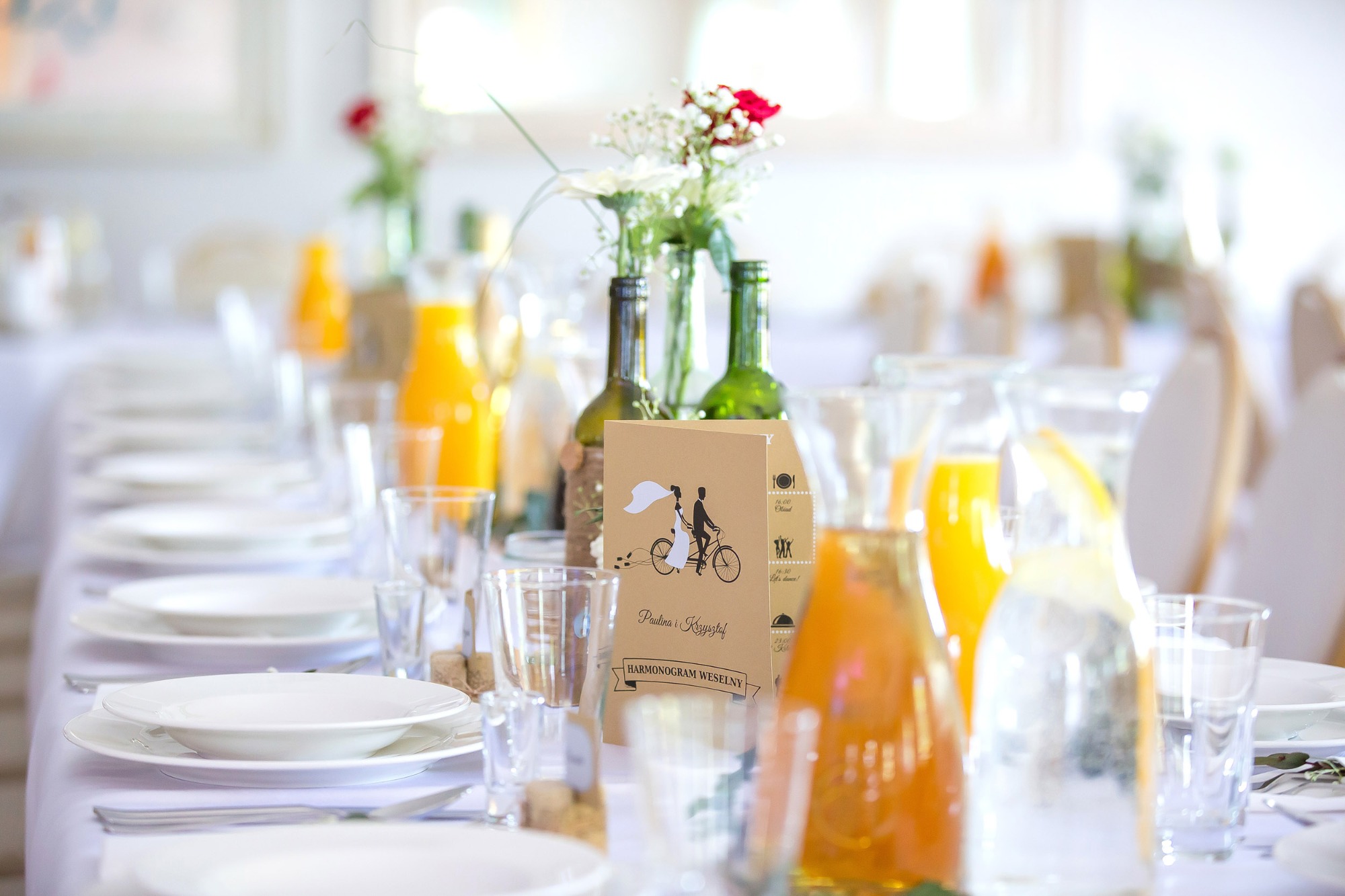Uroczysty stół weselny z białym obrusem, zastawą, dzbankami z sokiem, butelkami z kwiatami i kartą z harmonogramem wesela z motywem pary młodej na rowerze.