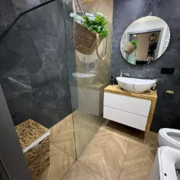 Nowoczesna łazienka z ciemnymi płytkami imitującymi kamień, drewnianą podłogą ułożoną w jodełkę i owalną umywalką nablatową. W rogu widoczny kosz wiklinowy, a nad prysznicem zawieszona doniczka...