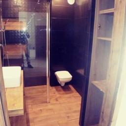 Wykończenia wnętrz kompleksowo - Nowoczesna łazienka z kabiną prysznicową typu walk-in, ciemnymi płytkami na ścianach, podłogą imitującą drewno i drewnianą zabudową meblową z otwartymi półkami.