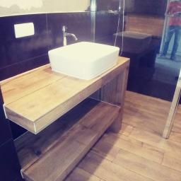 Wykończenia wnętrz kompleksowo - Nowoczesna łazienka z czarnymi płytkami i drewnianymi akcentami: umywalka nablatowa na drewnianym blacie z półkami, widoczna toaleta wisząca i kabina prysznicowa z odbiciem osoby.