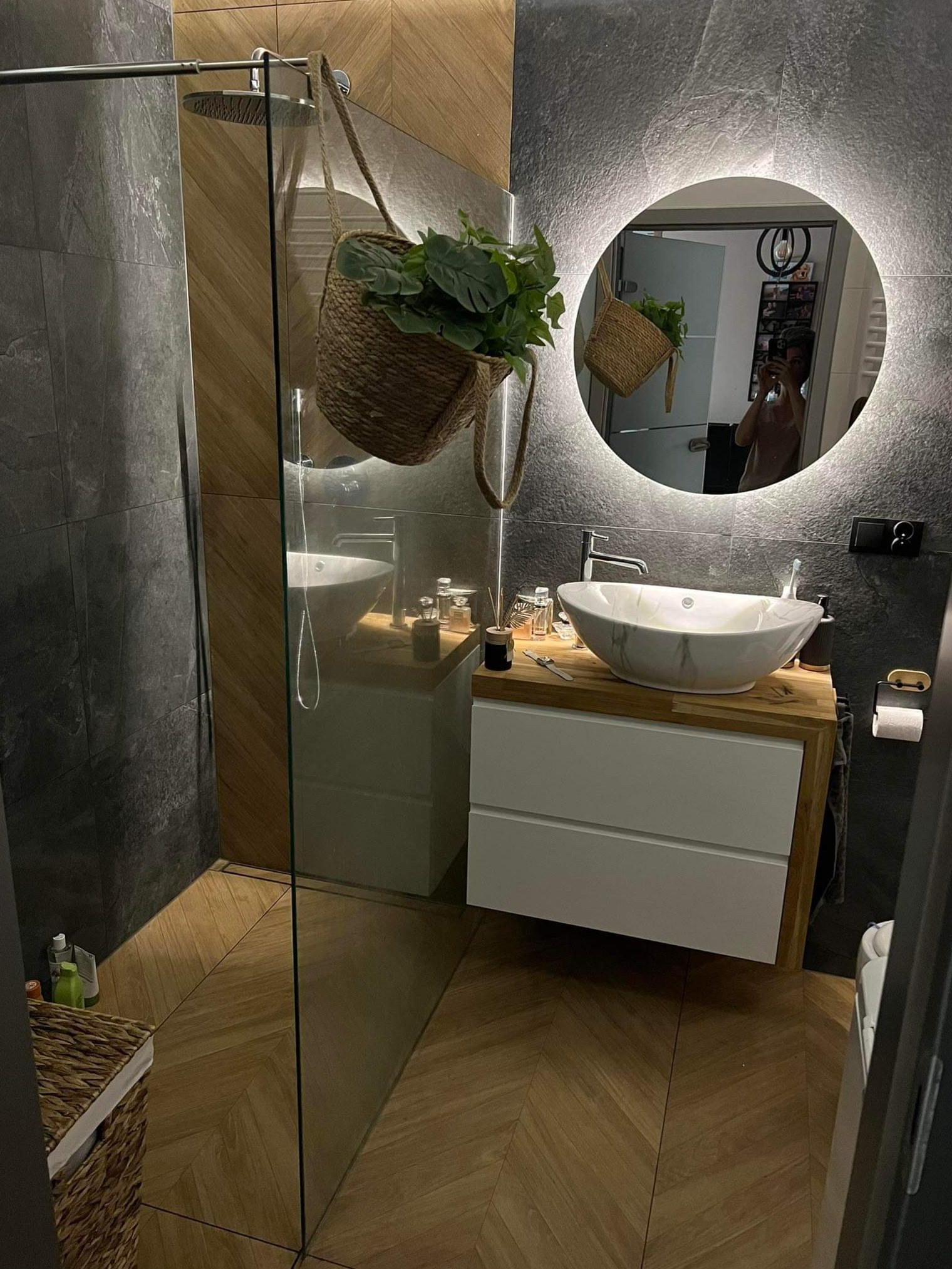 Nowoczesna łazienka z szarą płytką imitującą kamień, drewnianą podłogą w jodełkę, okrągłym lustrem z podświetleniem LED i umywalką nablatową z marmurowym wzorem.