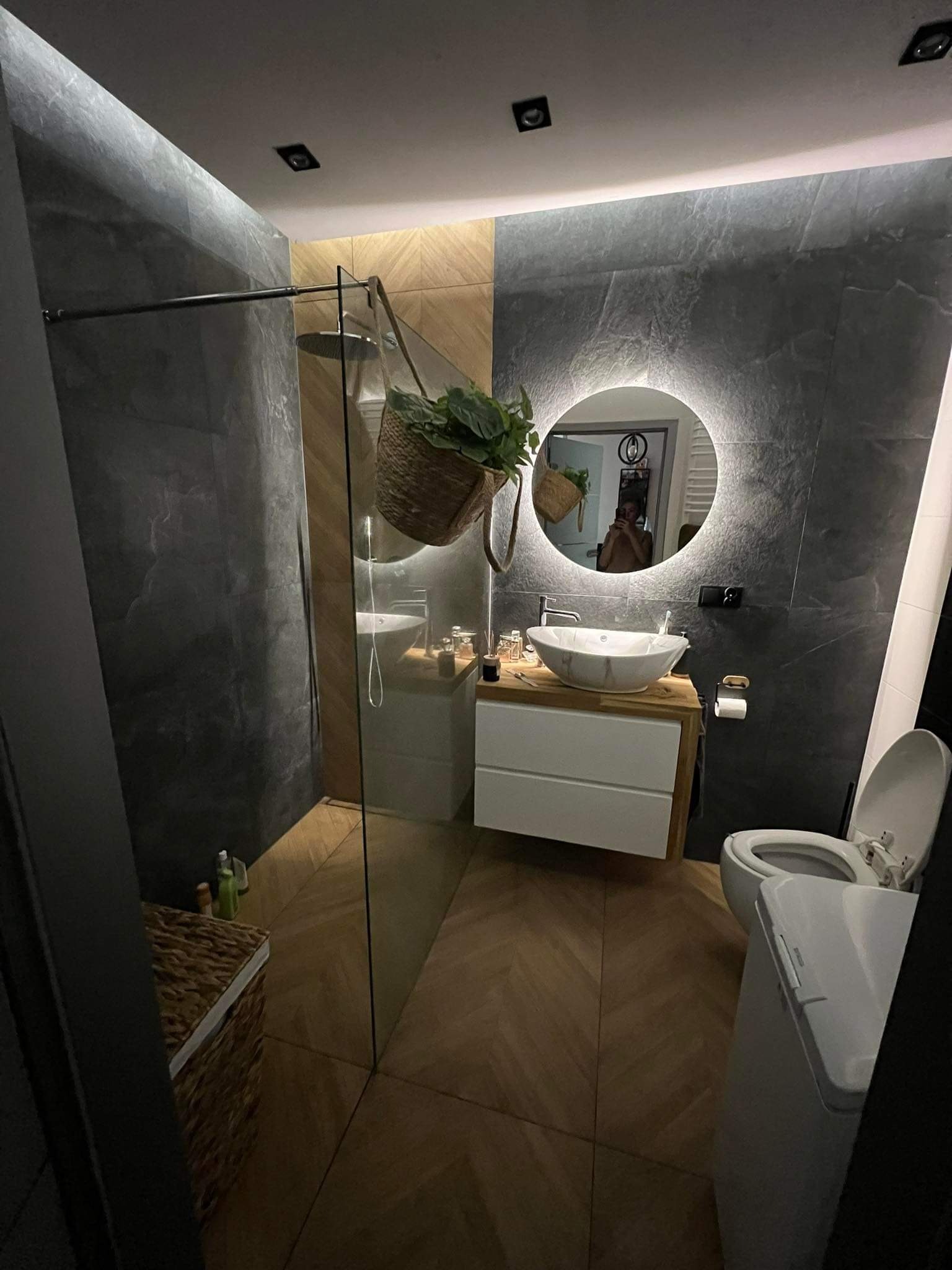 Nowoczesna łazienka z szarą, kamienną ścianą, okrągłym lustrem z podświetleniem LED, drewnianą podłogą w jodełkę i szklaną kabiną prysznicową typu walk-in, z wiszącym koszem z roślinami.
