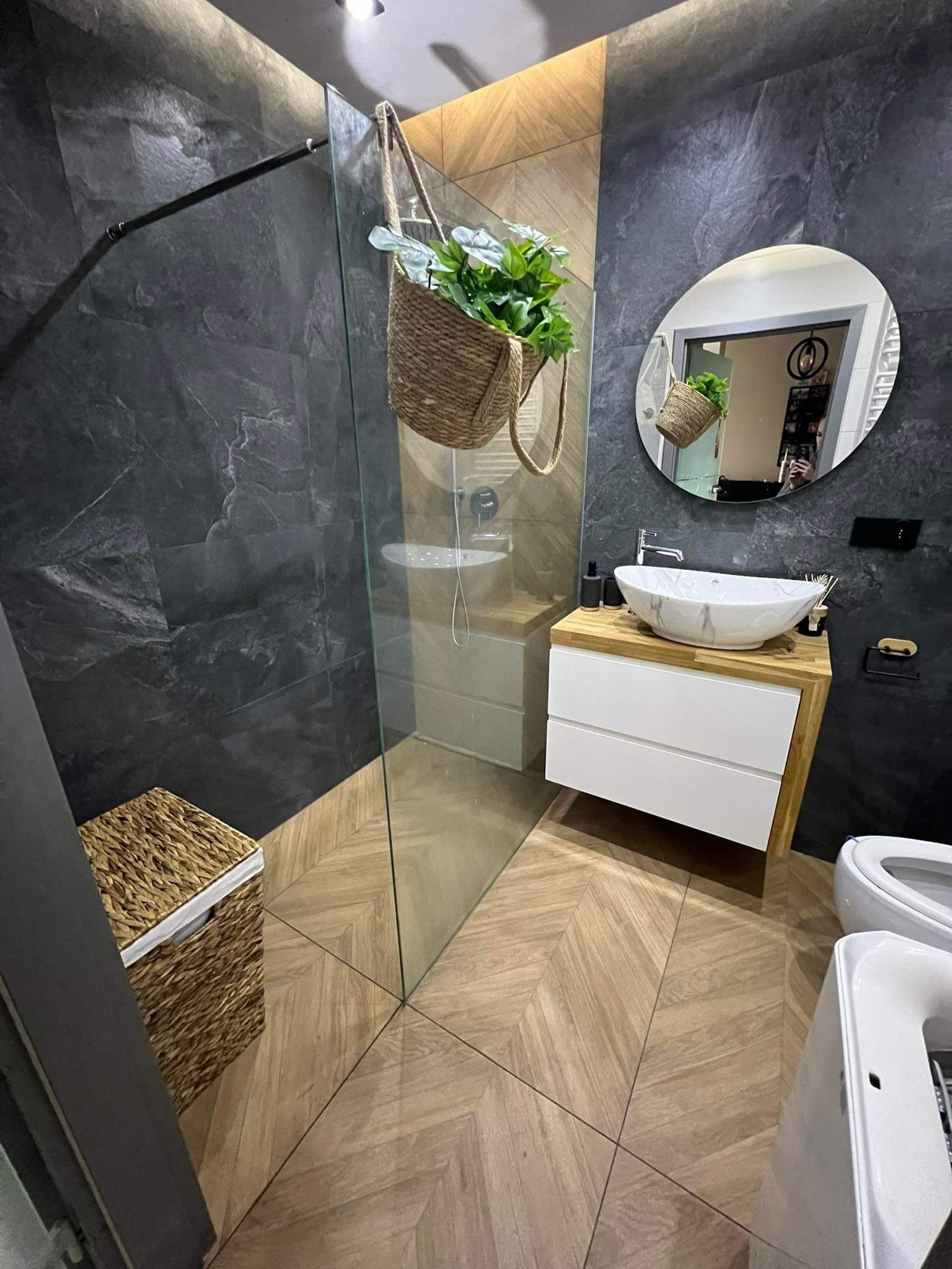 Nowoczesna łazienka z ciemnymi płytkami imitującymi kamień, drewnianą podłogą ułożoną w jodełkę i owalną umywalką nablatową. W rogu widoczny kosz wiklinowy, a nad prysznicem zawieszona doniczka...