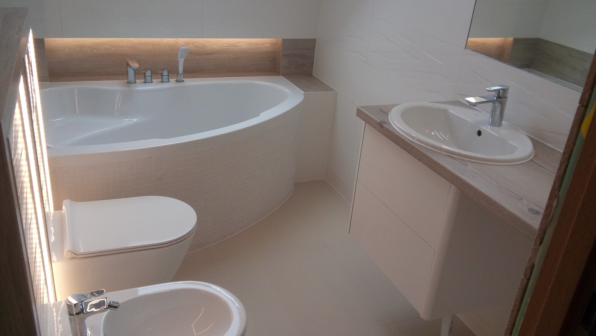 Nowoczesna, jasna łazienka z wanną obudowaną mozaiką, wiszącą umywalką z szafką o drewnianym blacie, toaletą i bidetem. Oświetlenie LED nad wanną. Minimalistyczny design.