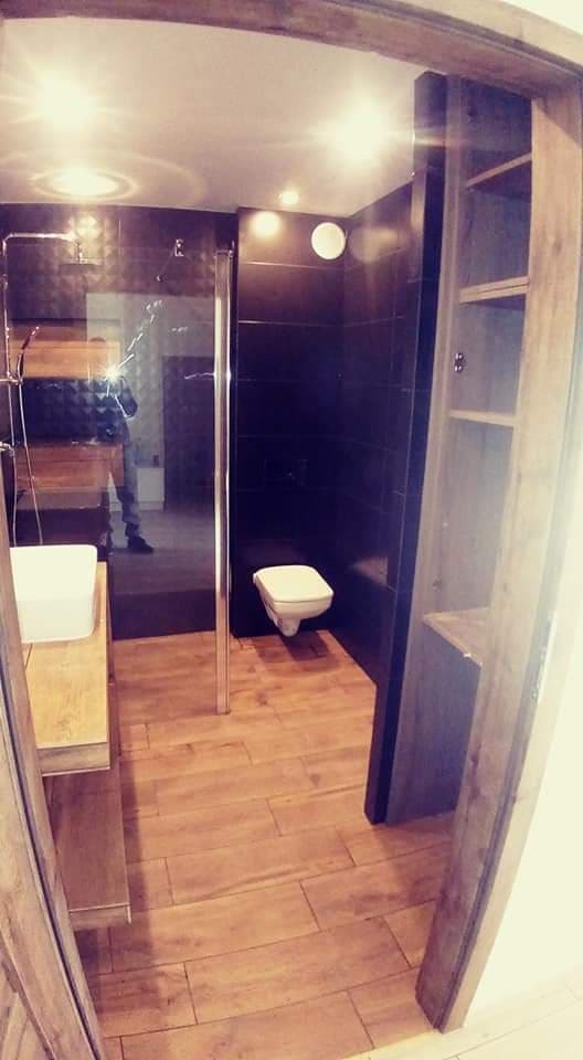 Nowoczesna łazienka z kabiną prysznicową typu walk-in, ciemnymi płytkami na ścianach, podłogą imitującą drewno i drewnianą zabudową meblową z otwartymi półkami.