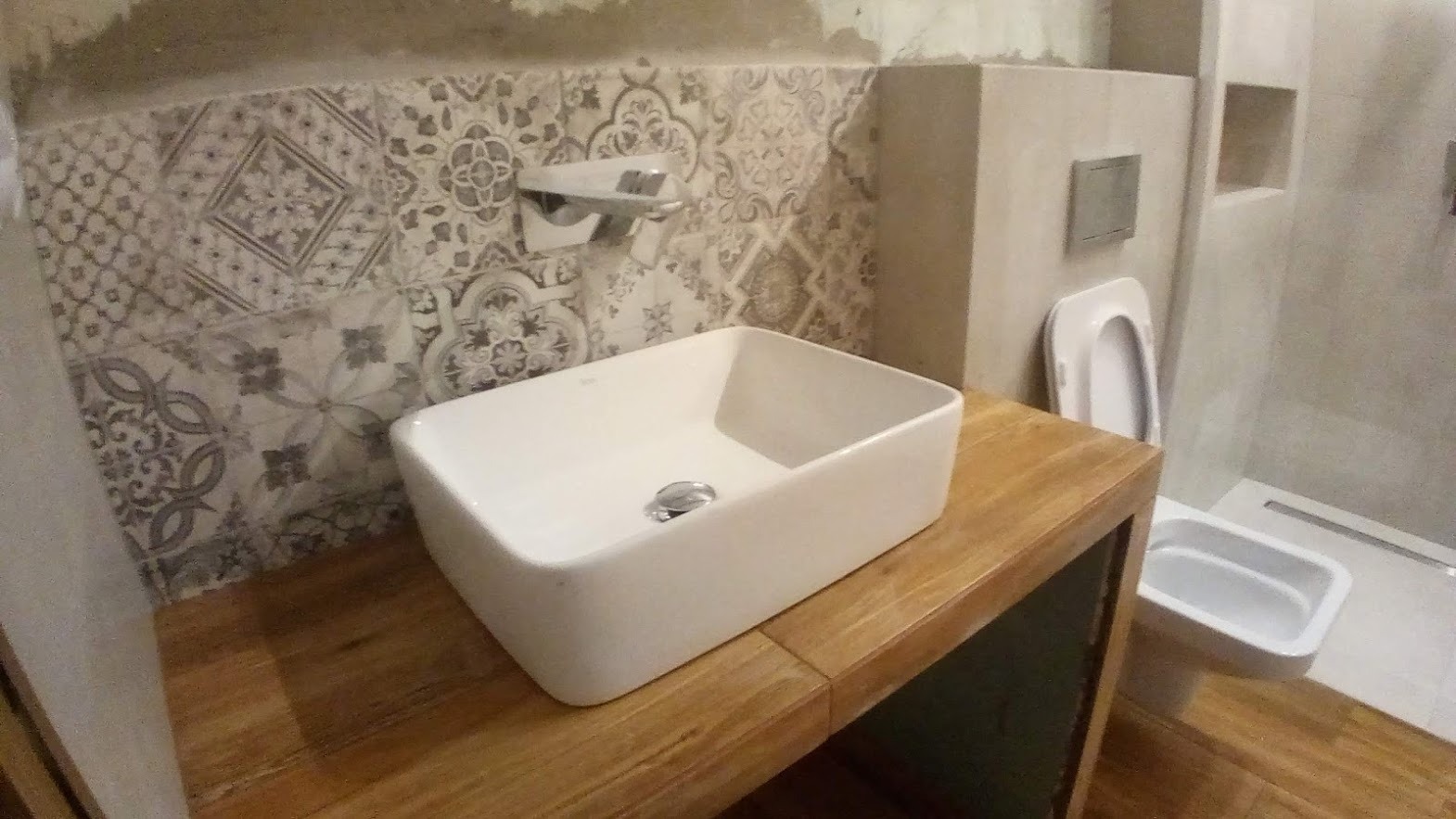 Nowoczesna łazienka z umywalką nablatową w kształcie prostokąta, drewnianym blatem, dekoracyjnymi płytkami na ścianie i podwieszaną toaletą z bidetem w tle.