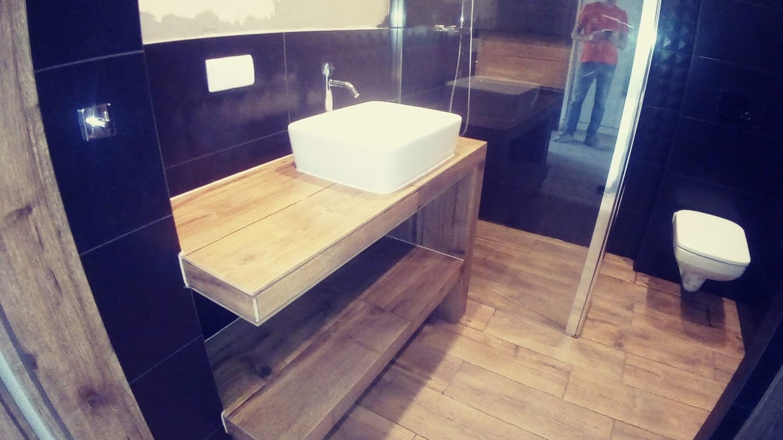 Nowoczesna łazienka z czarnymi płytkami i drewnianymi akcentami: umywalka nablatowa na drewnianym blacie z półkami, widoczna toaleta wisząca i kabina prysznicowa z odbiciem osoby.