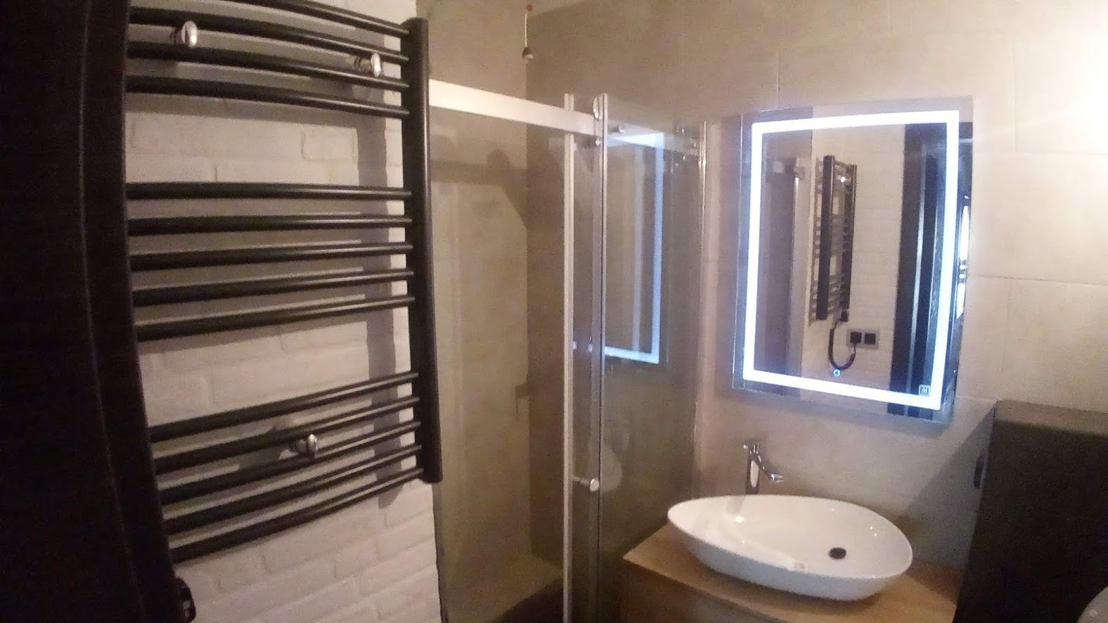 Nowoczesna łazienka z czarnym grzejnikiem drabinkowym na ścianie z białej cegły, szklaną kabiną prysznicową i podświetlanym lustrem nad białą umywalką.