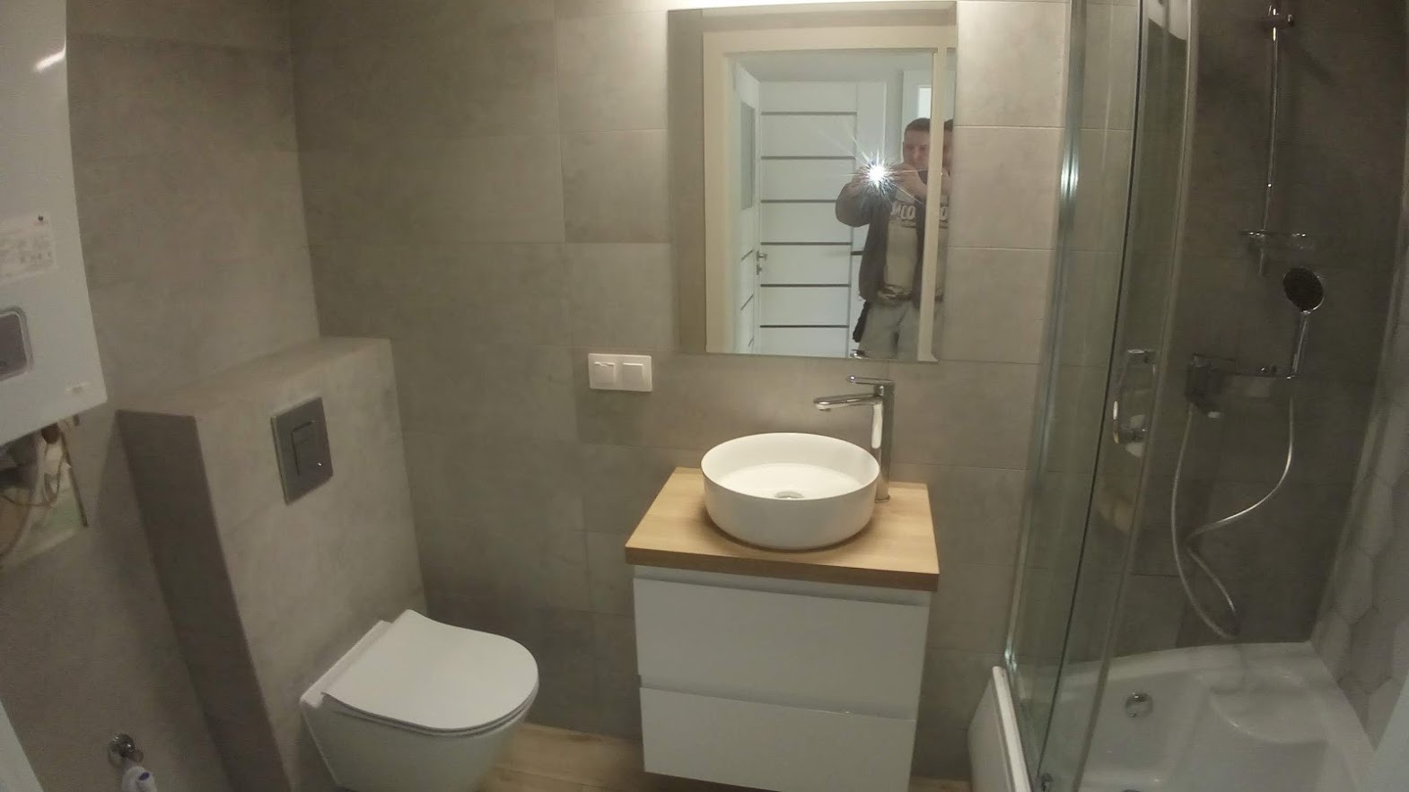 Nowoczesna łazienka w odcieniach szarości z kabiną prysznicową, umywalką nablatową na drewnianym blacie i wiszącą toaletą; widoczne odbicie fotografa w lustrze.