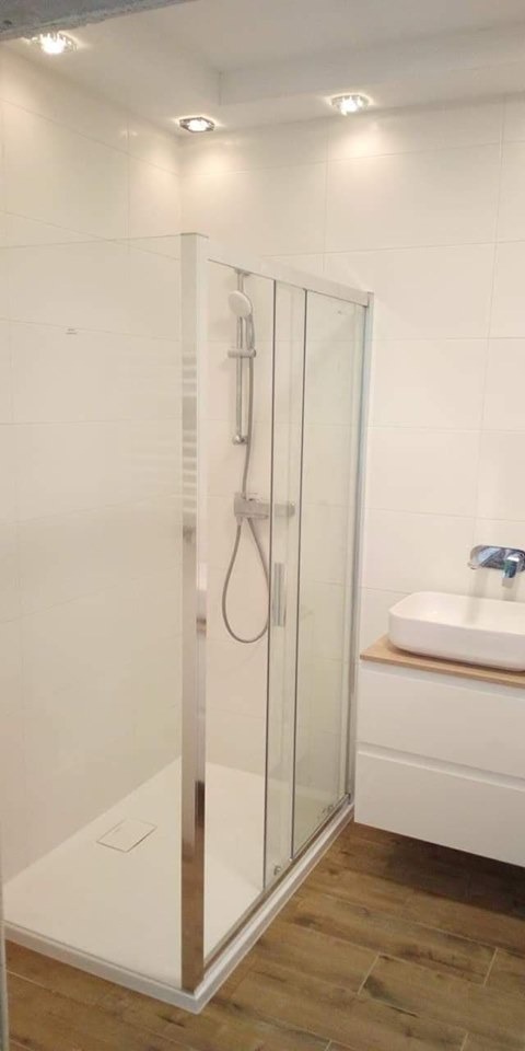 Nowoczesna łazienka z kabiną prysznicową ze srebrnymi profilami, białą umywalką nablatową i drewnopodobną podłogą z płytek.