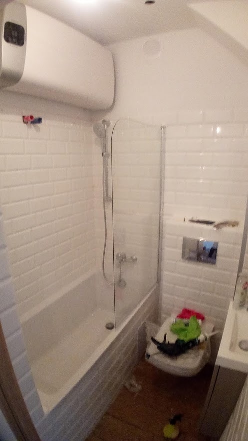 Nieduża łazienka po remoncie z białą wanną z parawanem, białą cegiełkową glazurą na ścianach i drewnianą podłogą. Na sedesie leżą ubrania.