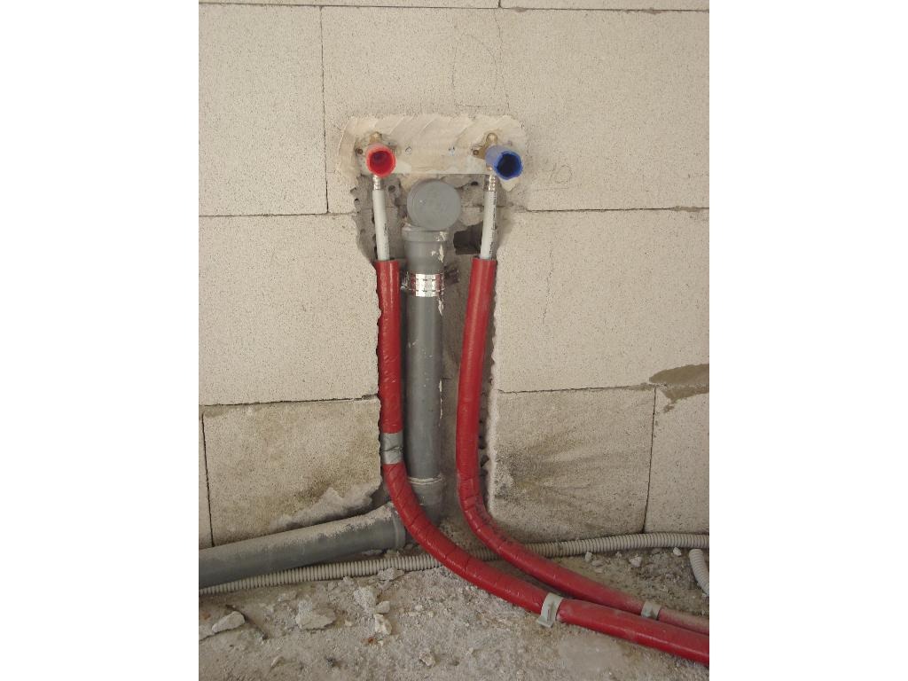 Wewnętrzna instalacja wodno-kanalizacyjna w surowej łazience, widoczne rury ciepłej i zimnej wody z izolacją, szare rury odpływowe i betonowe bloczki ściany.