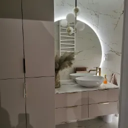 Nowoczesna łazienka z podwójną umywalką nablatową, szafką z szufladami w kolorze pudrowego różu, okrągłym lustrem z podświetleniem LED i marmurowymi płytkami na ścianie.