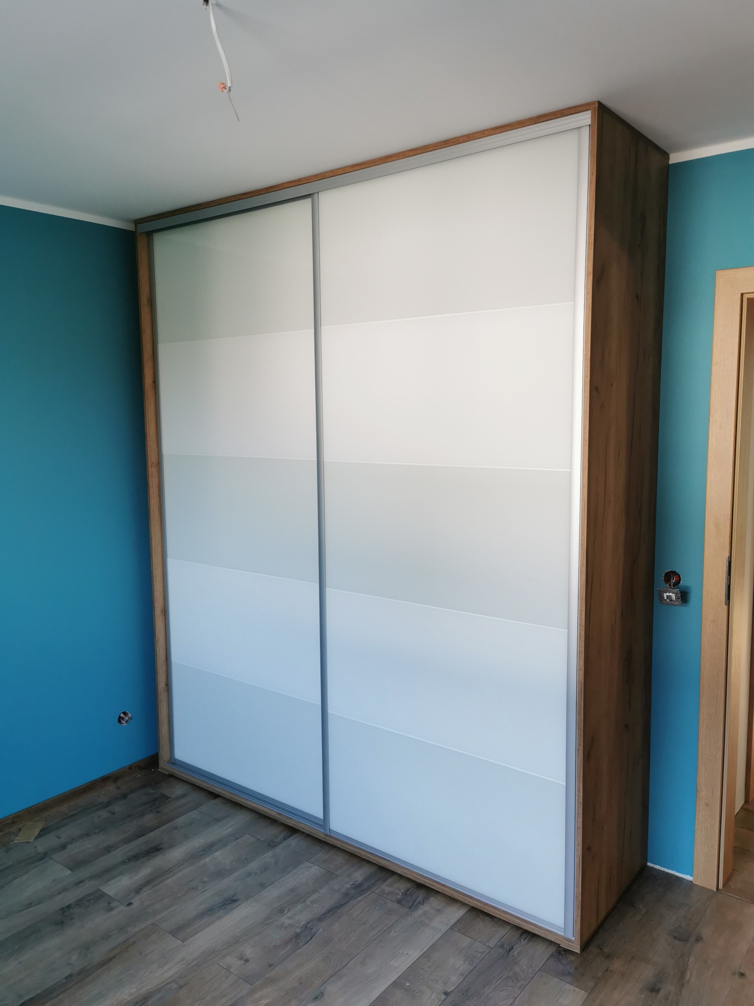Szafa wnękowa z przesuwnymi drzwiami w odcieniach bieli i szarości, drewniane wykończenie, turkusowa ściana w tle.