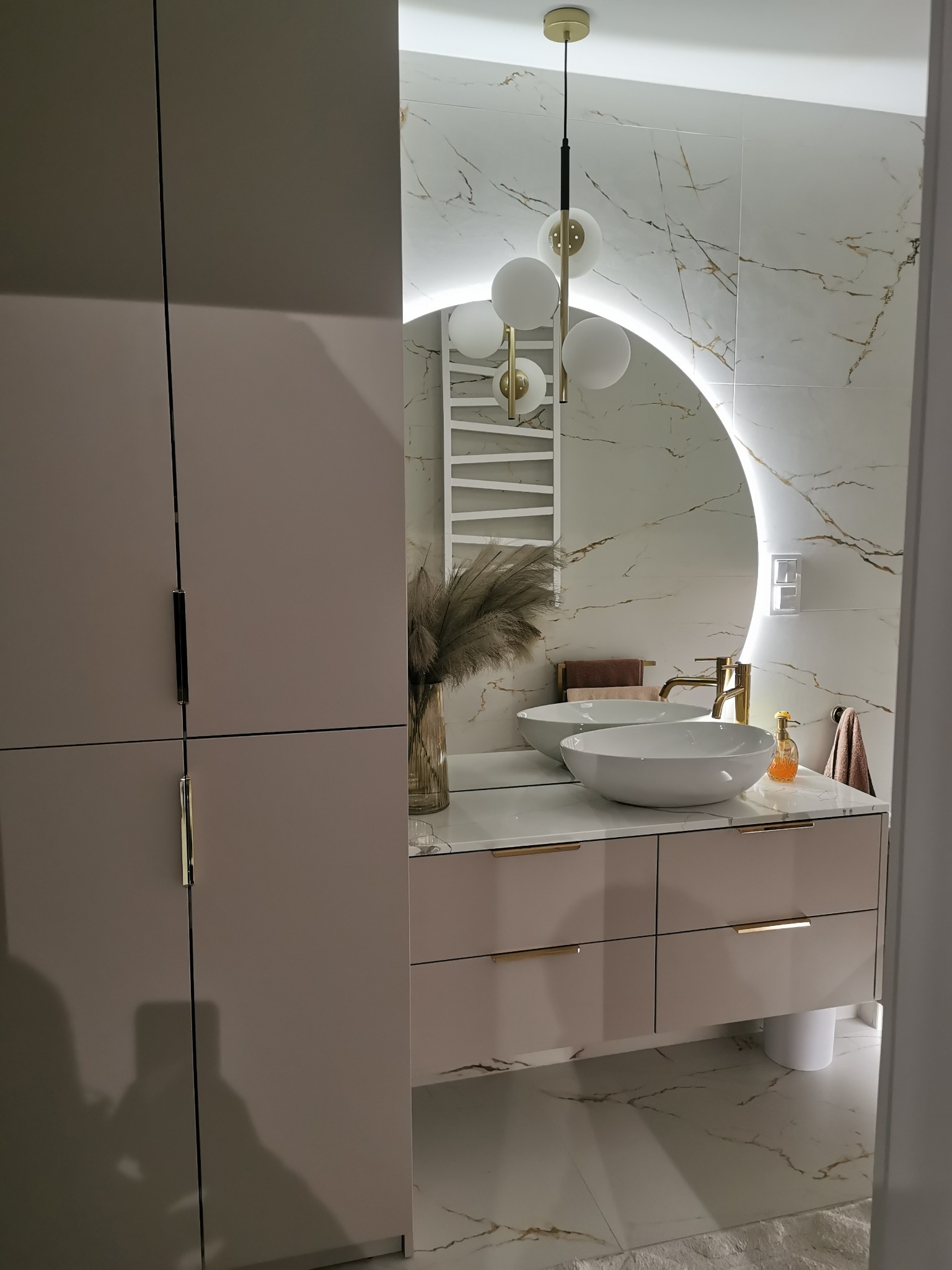 Nowoczesna łazienka z podwójną umywalką nablatową, szafką z szufladami w kolorze pudrowego różu, okrągłym lustrem z podświetleniem LED i marmurowymi płytkami na ścianie.