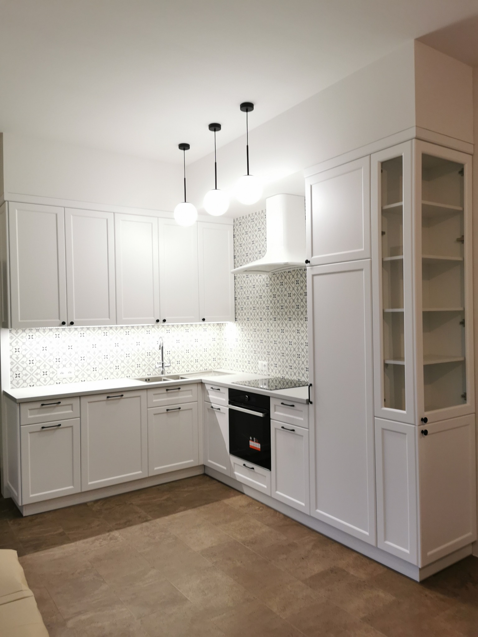 Jasna kuchnia z białymi szafkami, wzorzystą mozaiką nad blatem i trzema wiszącymi lampami kulami nad zlewem, widziana z niskiego kąta.