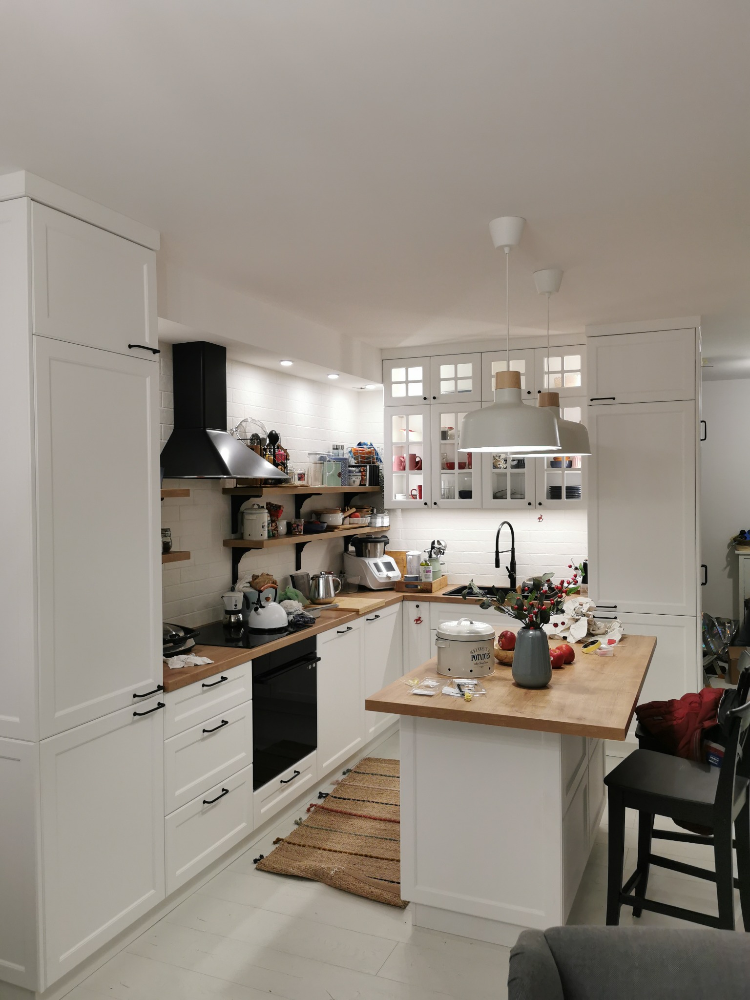 Jasna kuchnia z białą zabudową, drewnianym blatem i czarnymi akcentami, widok na wyspę kuchenną z akcesoriami i elementami dekoracyjnymi.