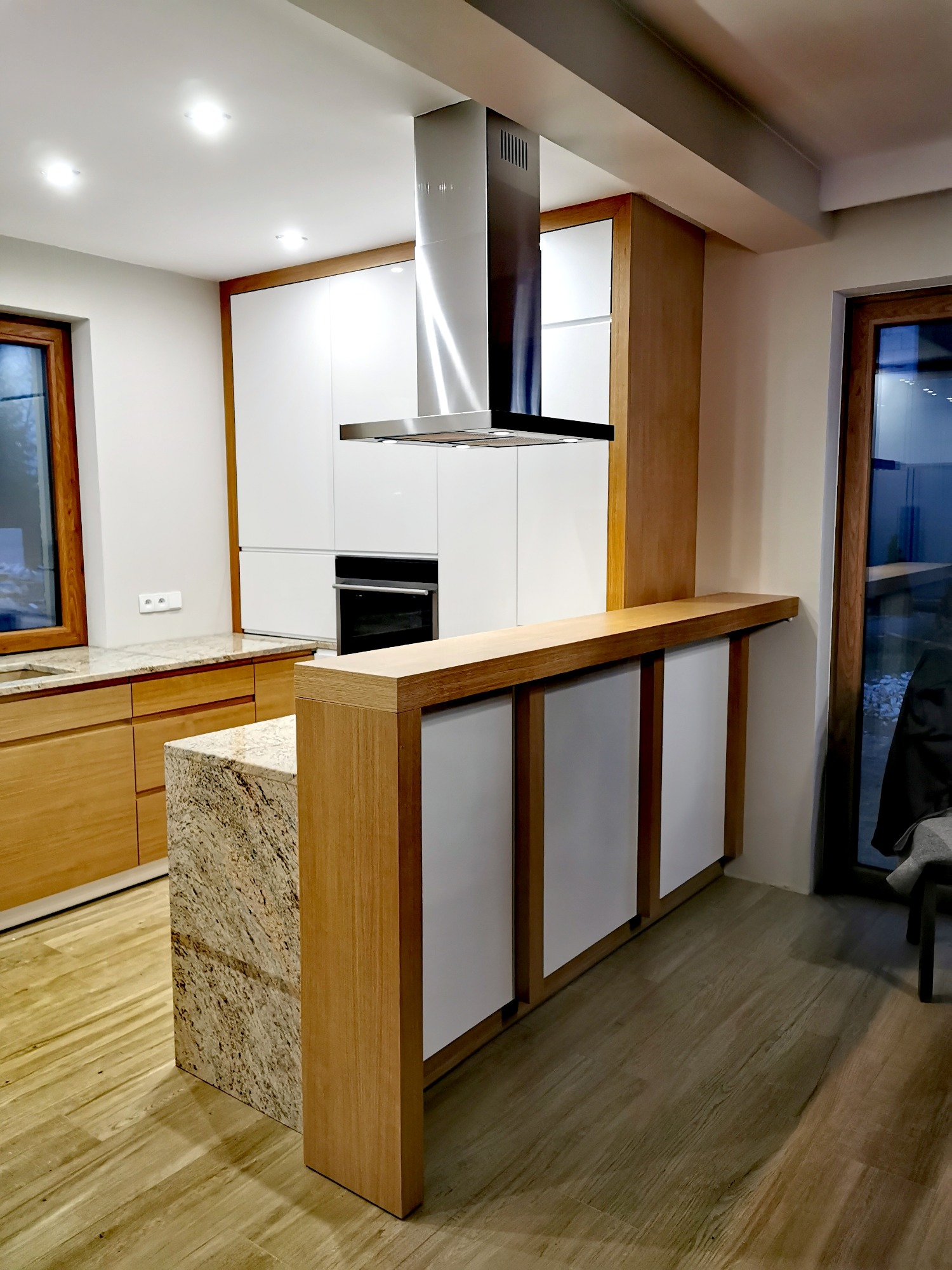 Nowoczesna kuchnia z wyspą wykończoną kamiennym blatem, drewnianymi elementami i białymi frontami szafek, oświetlona punktowymi lampami sufitowymi.