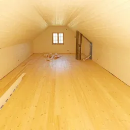 Poddasze w trakcie adaptacji: świeżo ułożona, jasna drewniana podłoga z widocznymi belkami stropowymi i oknem. Część desek podłogowych jeszcze nieułożona leży obok ściany.