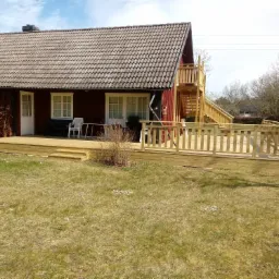 Czerwony dom z drewnianym tarasem, balustradą i schodami prowadzącymi na górę, otoczony trawnikiem z widocznymi stokrotkami.