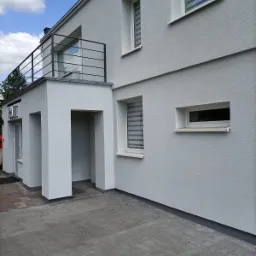 Jasnoszary dom z wykończoną elewacją, balkonem z czarną balustradą i szarym brukowanym podjazdem, widok z zewnątrz w słoneczny dzień.