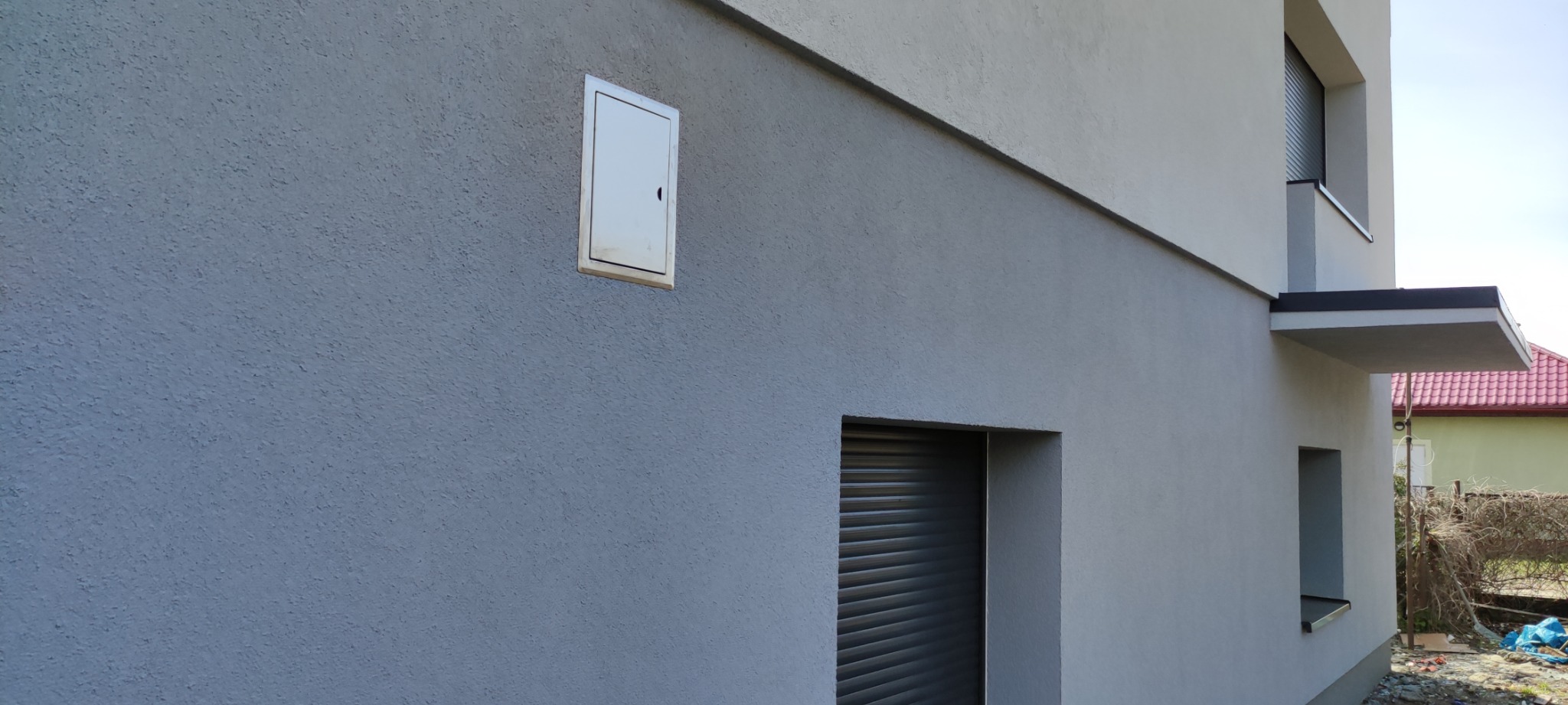 Dwukolorowa elewacja budynku z szarą, chropowatą teksturą tynku i wbudowaną, metalową skrzynką rewizyjną oraz oknem z opuszczoną roletą zewnętrzną.