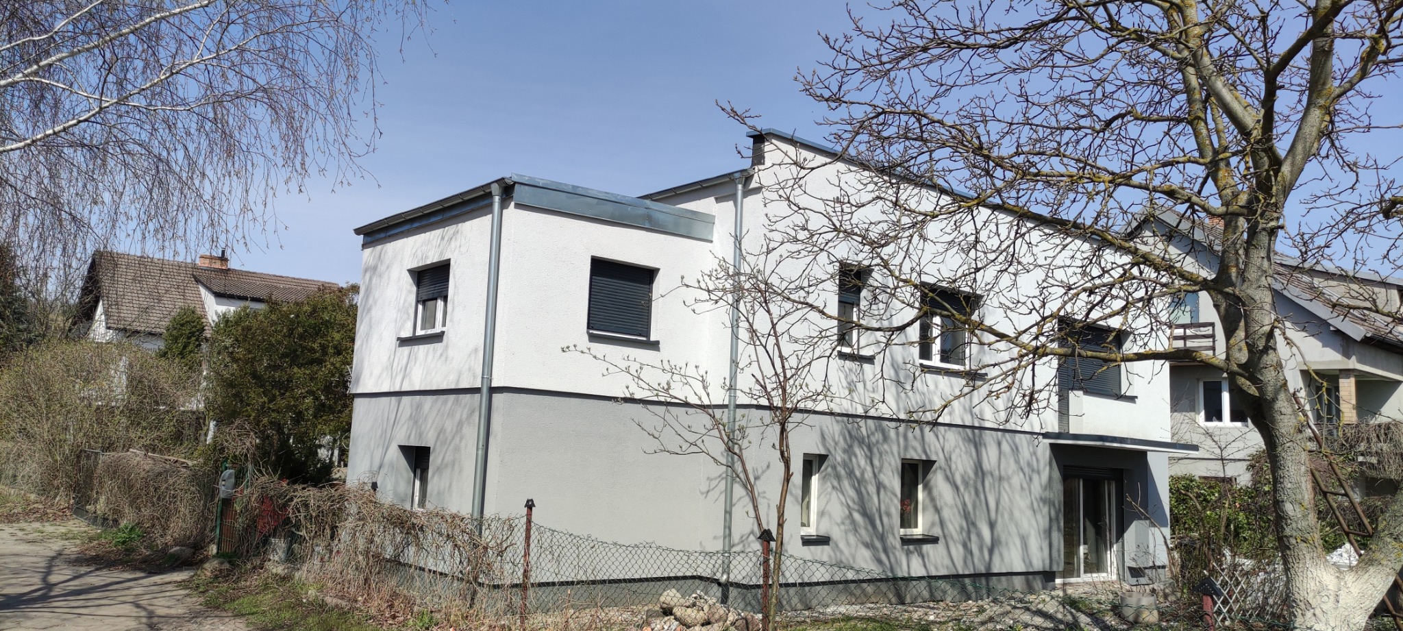 Dwukondygnacyjny dom z elewacją w odcieniach szarości, z roletami zewnętrznymi w oknach, widziany z perspektywy ogrodu z drzewami bezlistnymi na pierwszym planie.