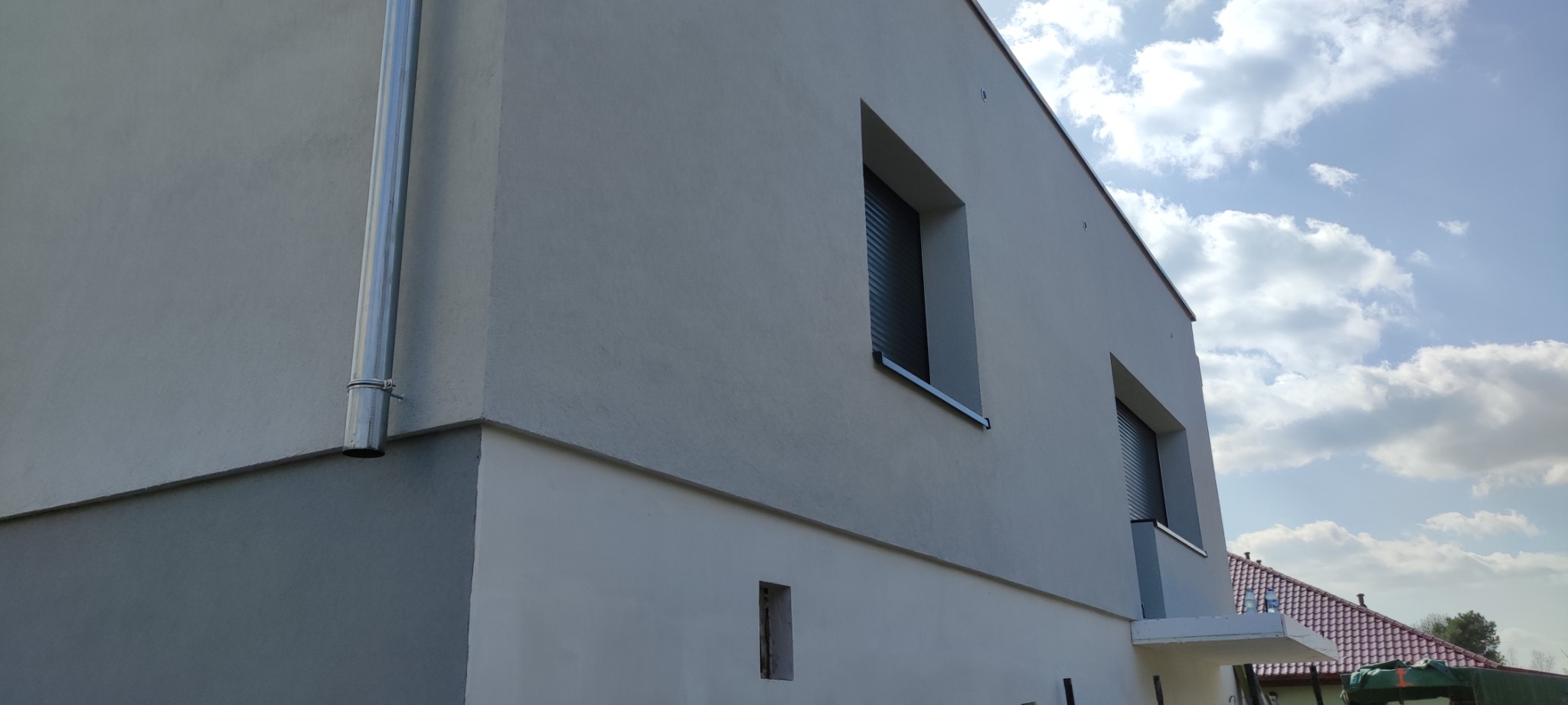 Dwukondygnacyjny budynek o jasnej elewacji z widocznym systemem rynnowym i oknami z roletami zewnętrznymi, sfotografowany pod kątem z częściowym widokiem nieba z chmurami.