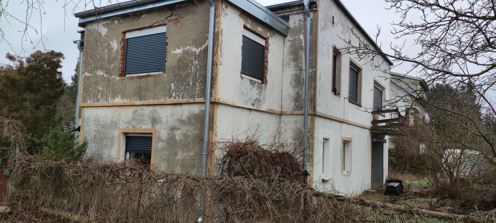 Dwukondygnacyjny dom z częściowo zniszczoną elewacją, widoczne ślady odpadającego tynku i uszkodzenia muru, okna z roletami zewnętrznymi, rynny wzdłuż krawędzi dachu, zaniedbany ogród z krzewami...