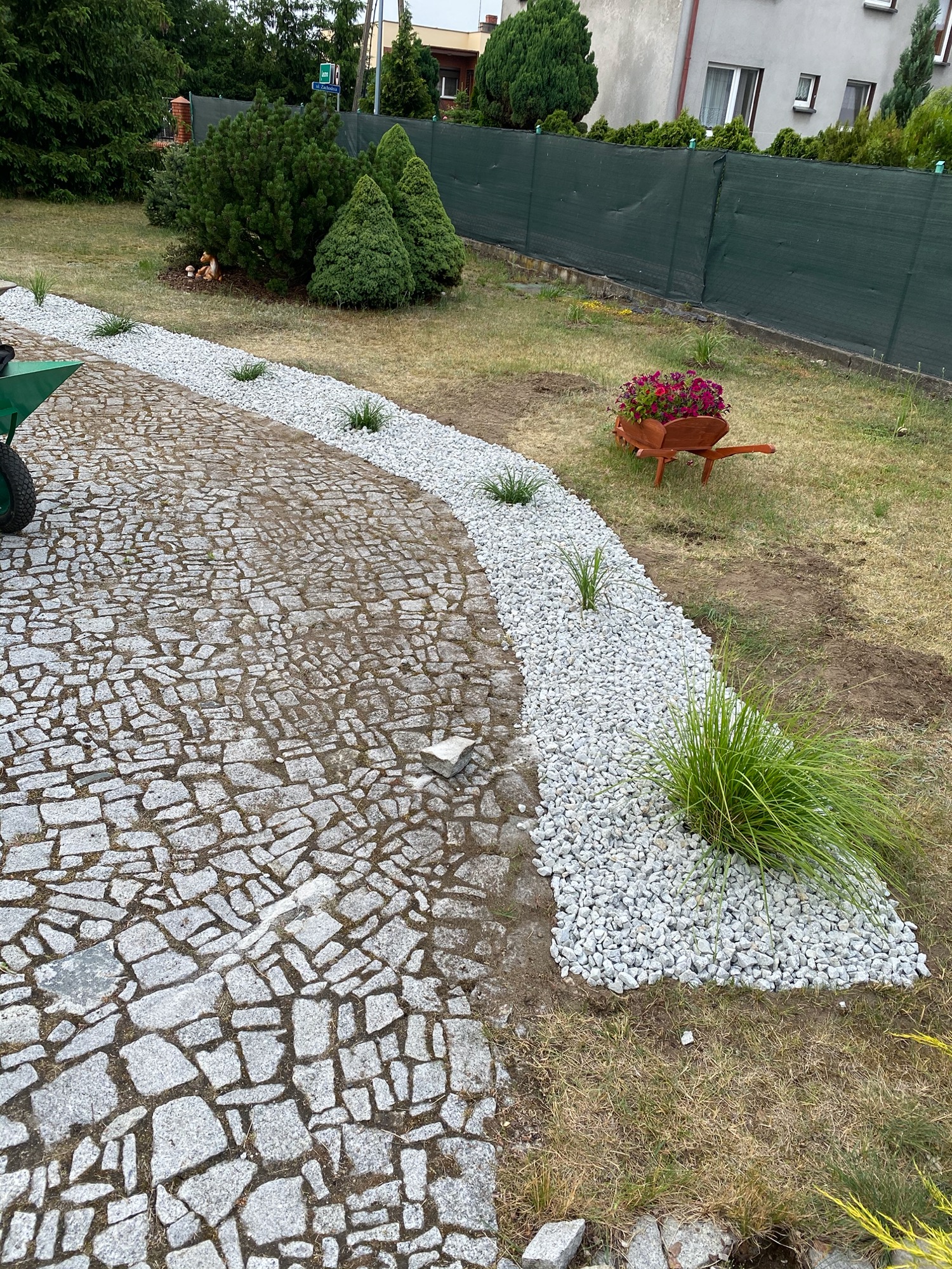 Podjazd z kostki granitowej obrzeżony pasem białego grysu z nasadzeniami ozdobnych traw, w tle drewniana taczka z kwiatami i zielony płot.