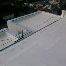 Widok z góry na płaski dach budynku pokryty białą membraną z widocznymi obróbkami blacharskimi i elementami instalacji odgromowej.