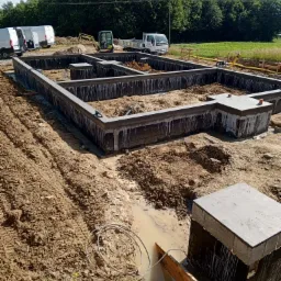 Wykop pod fundamenty domu jednorodzinnego z widocznymi zbrojeniami, deskowaniem i izolacją przeciwwilgociową, na działce budowlanej z zaparkowanymi pojazdami i drzewami w tle.